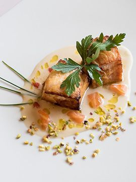 Pesce scottato servito su un piatto bianco con guarnizione di erbe aromatiche, salsa a pezzetti e briciole sparse.