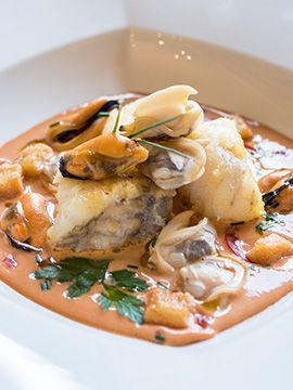 Filetto di pesce servito su un piatto bianco, con vongole e salsa, guarnito con prezzemolo.