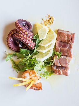 Polpo, carne a fette, spicchi di limone e guarnizione disposti su un piatto bianco.