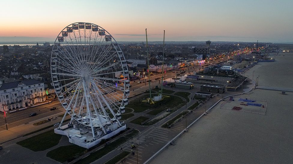 Ferris wheel on a beach promenade at dusk with buildings, road, and ocean in view.