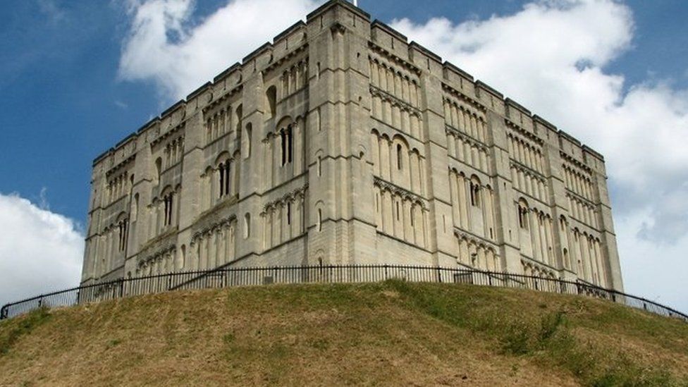Square stone castle on a grassy mound under a blue sky with clouds.