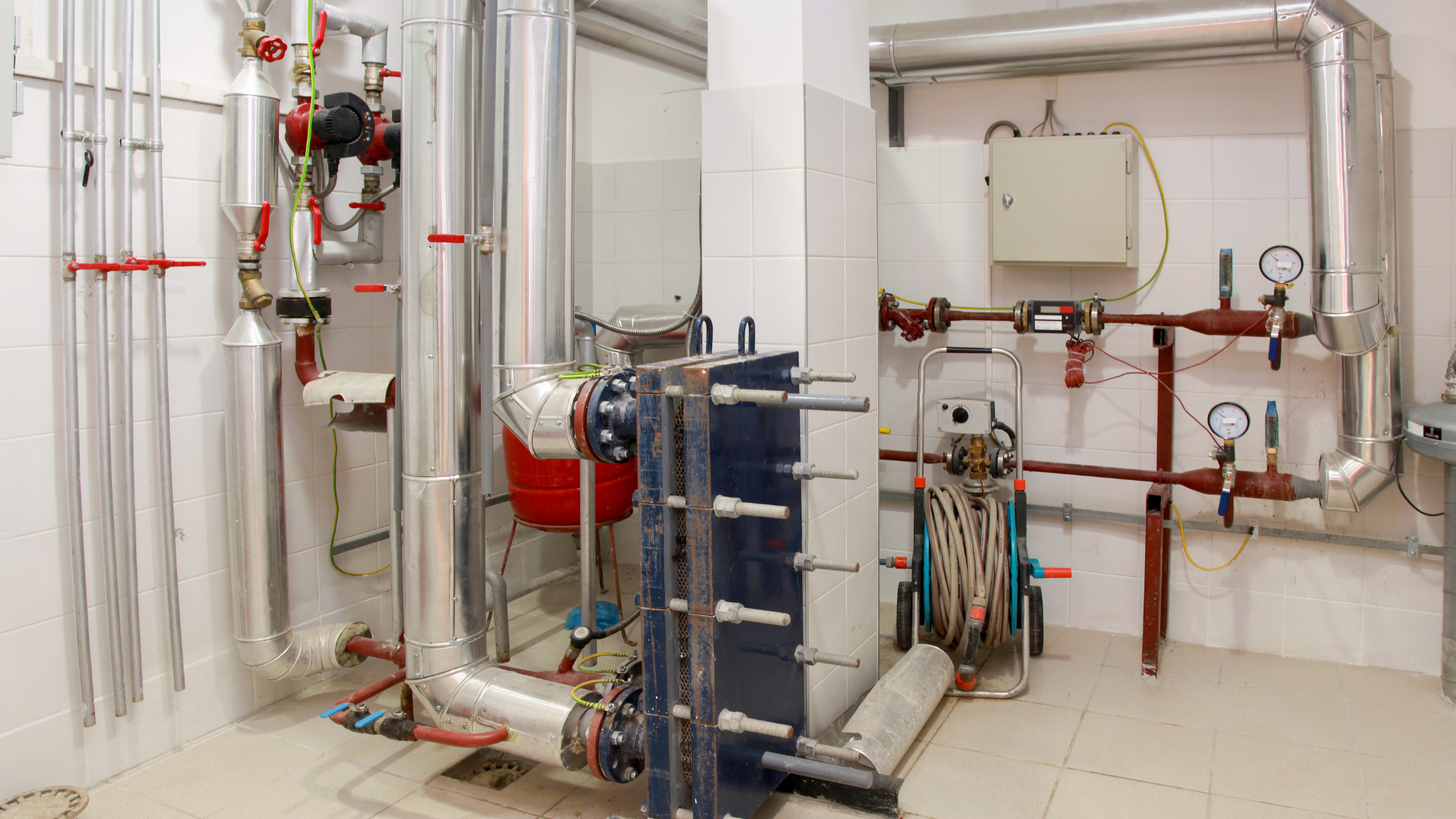 A room with industrial plumbing, pipes, valves, and a red pressure tank.