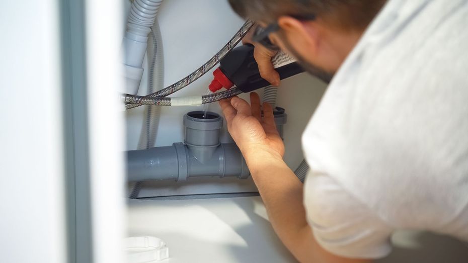 Person using a red drill to work on pipes under a sink, seen from behind.