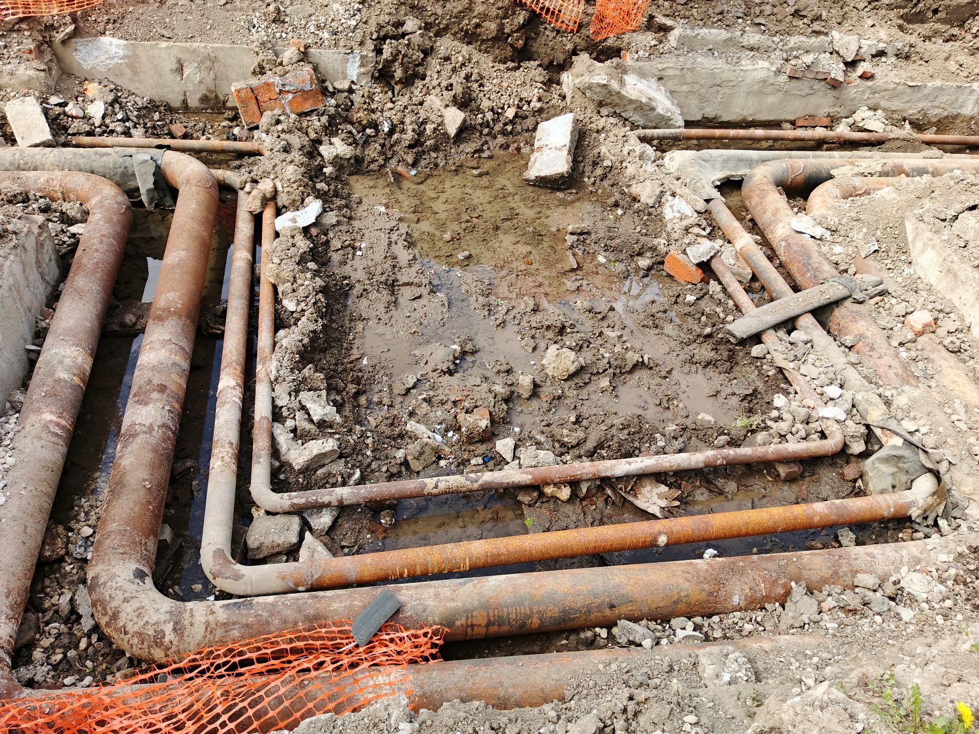 Exposed rusty pipes and muddy trench at a construction site with broken concrete and rubble