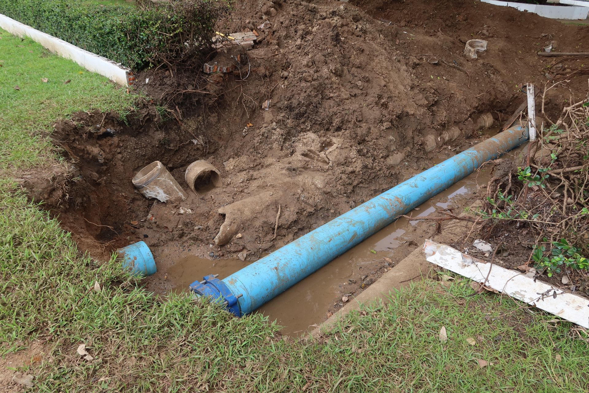 Broken blue pipe leaking water in a muddy ditch beside grass.