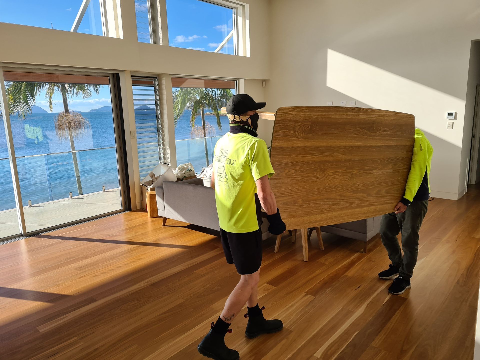 Two movers carrying a large wooden table through a sunlit room with ocean views — MM Removals In Nelson Bay, NSW