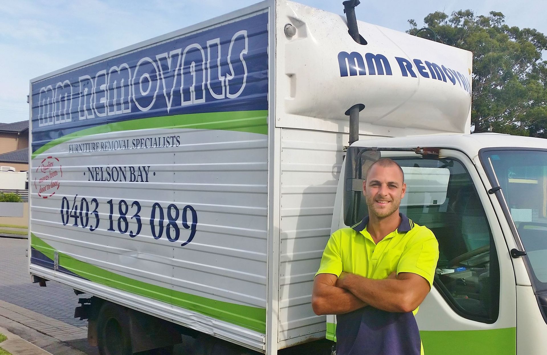 Man Posing Beside a White and Green Moving Truck with Company Branding — MM Removals In Nelson Bay, NSW