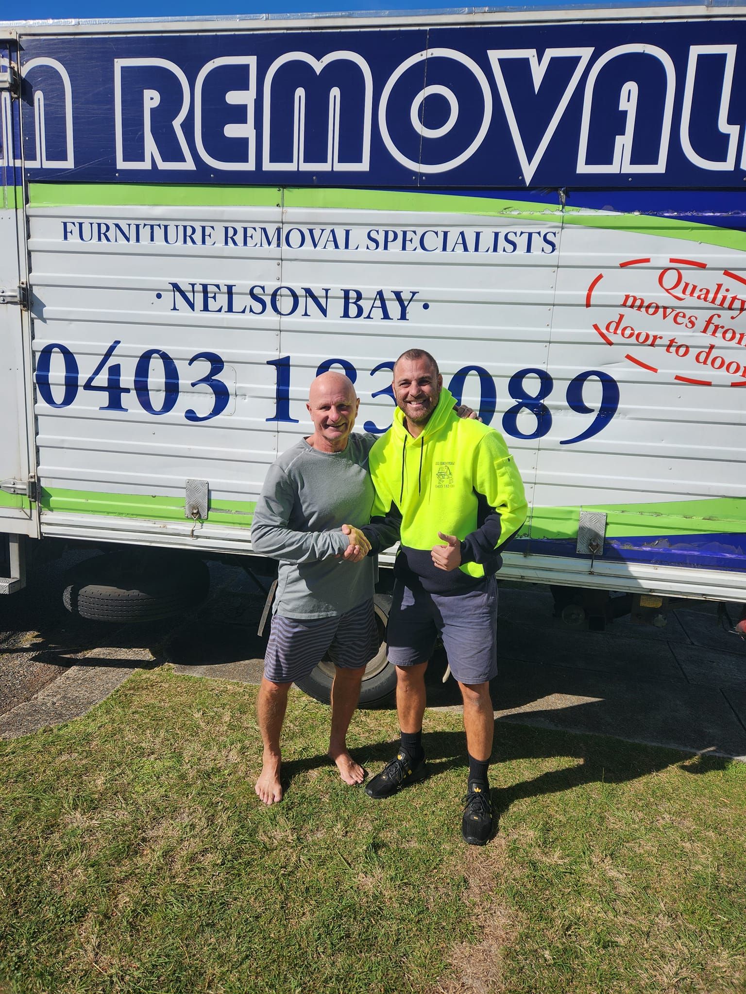 Two People Standing in Front of A Removal Truck, One in A Neon Yellow Shirt, Holding a Sign — MM Removals In Nelson Bay, NSW