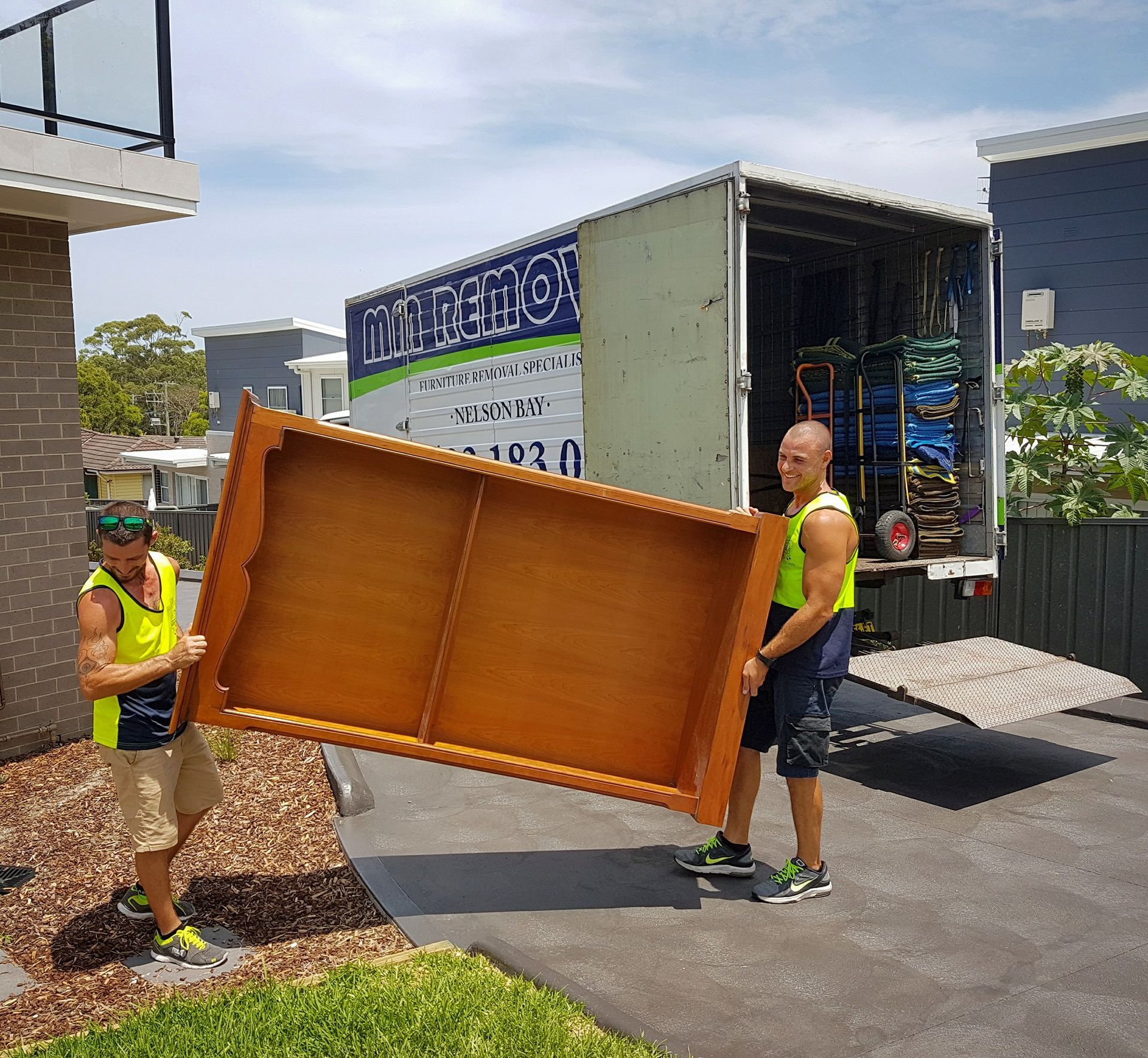 Two Workers in Neon Vests Carrying a Large Wooden Panel Outside a Building — MM Removals In Nelson Bay, NSW