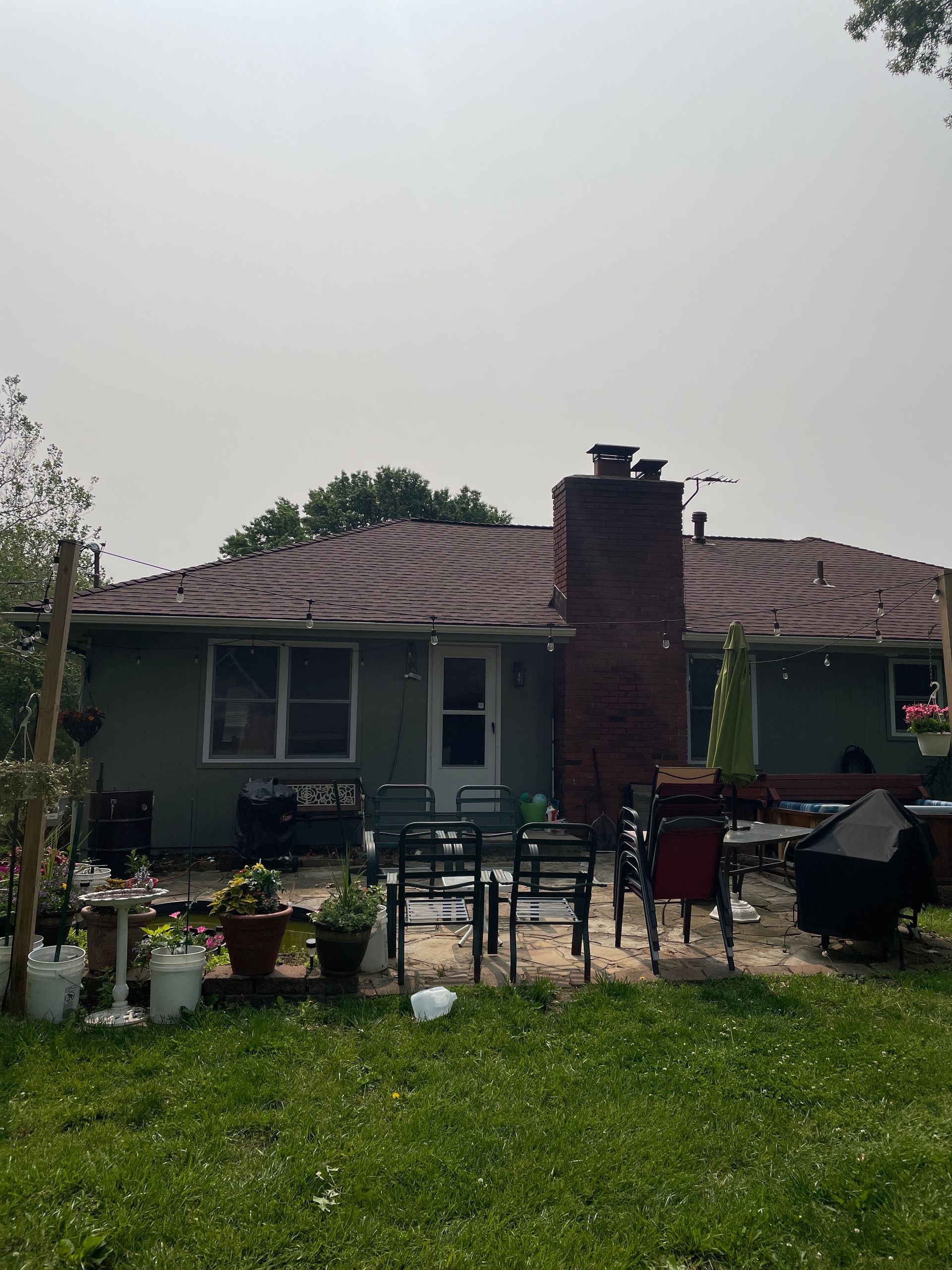 Backyard view of a single-story house with a brick chimney, patio furniture, and potted plants on a green lawn