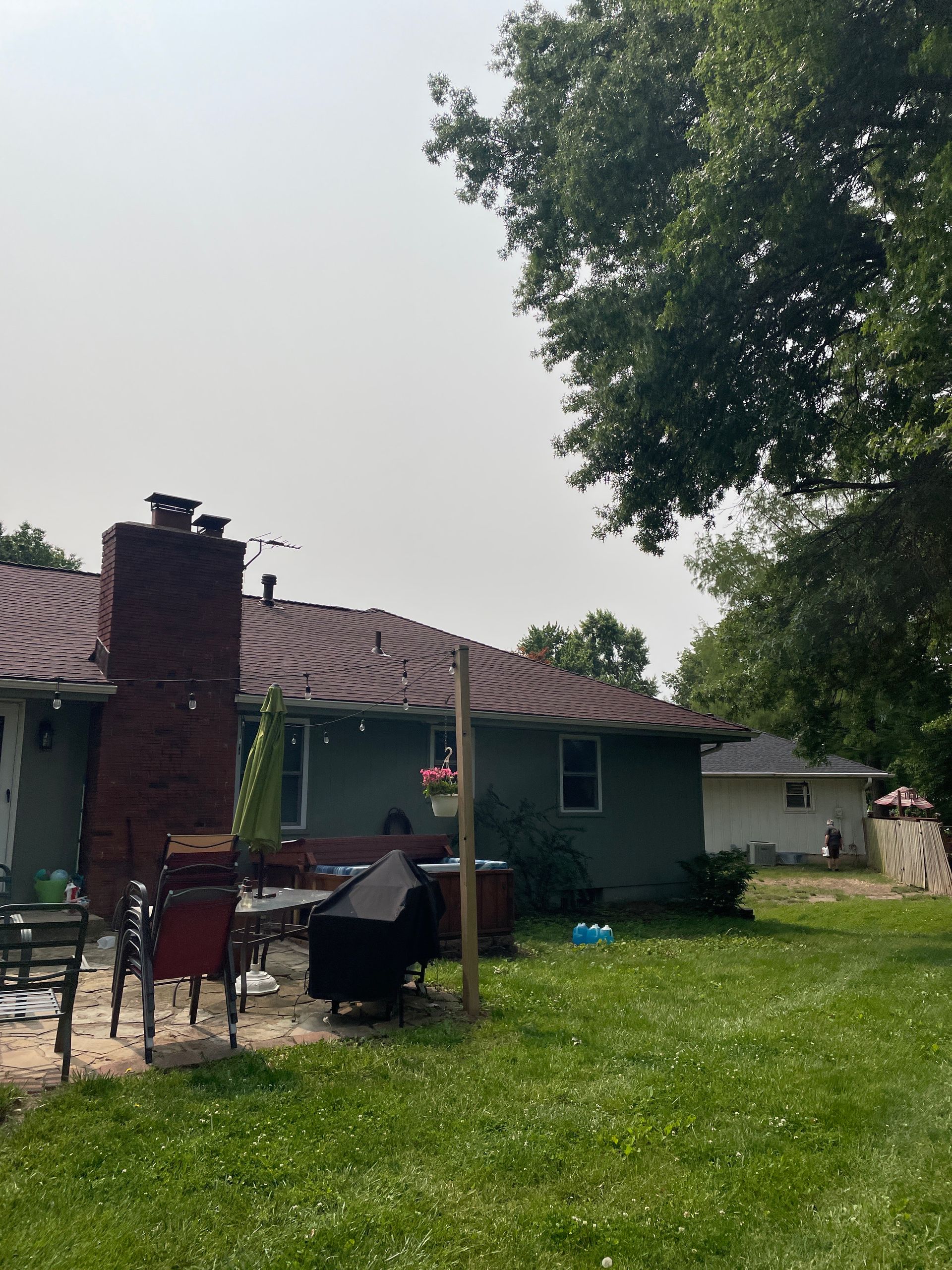 Backyard patio with a brick grill, chairs, a small house, and a large tree on a sunny day