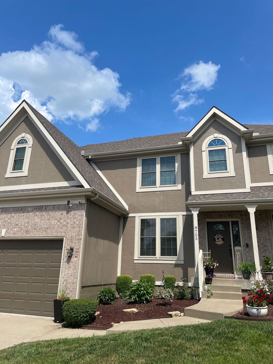 Suburban two-story beige house with garage, front porch, and landscaped yard under a blue sky.