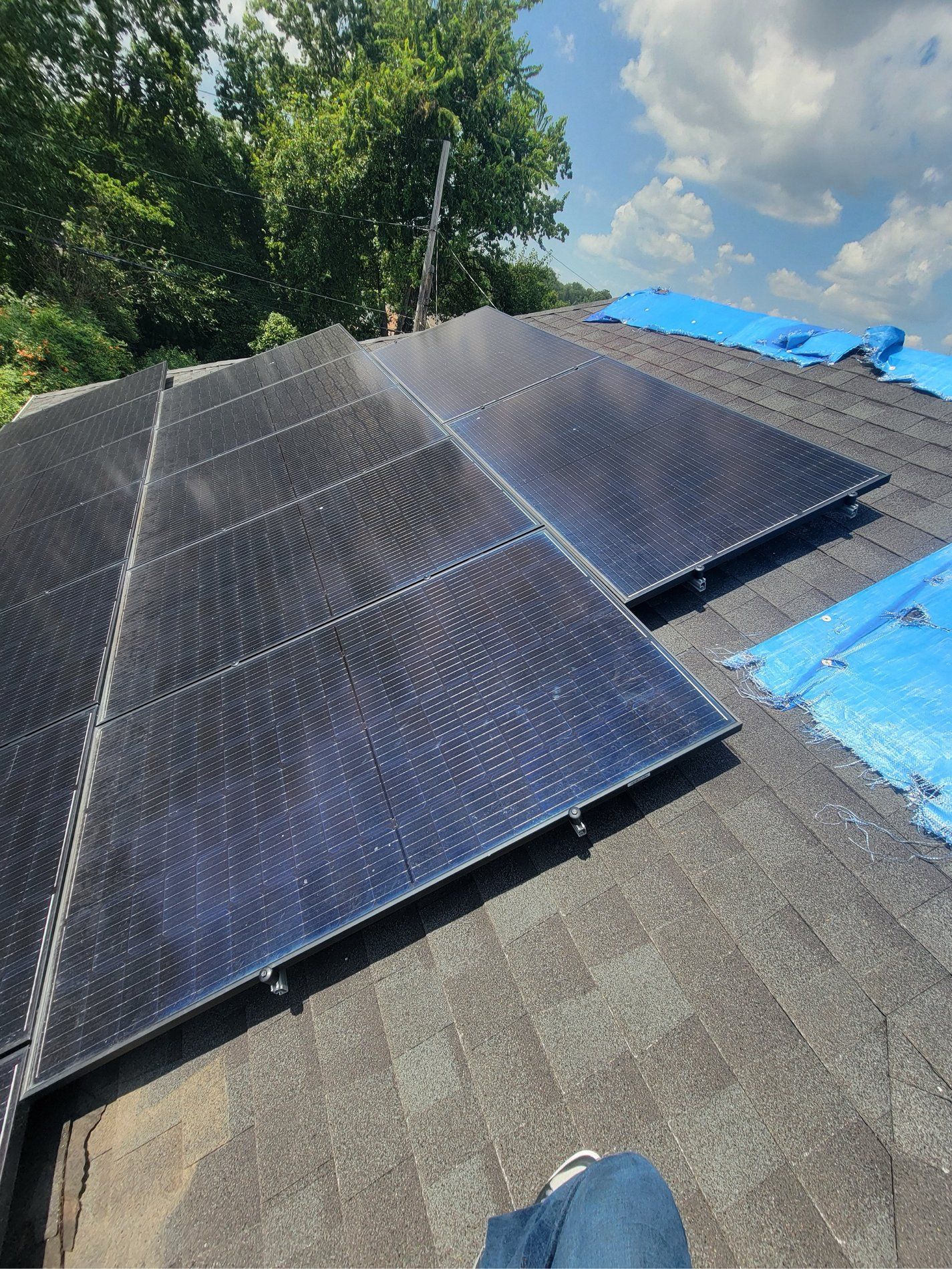 Solar panels installed on a roof beside a blue tarp, with trees and cloudy sky in the background
