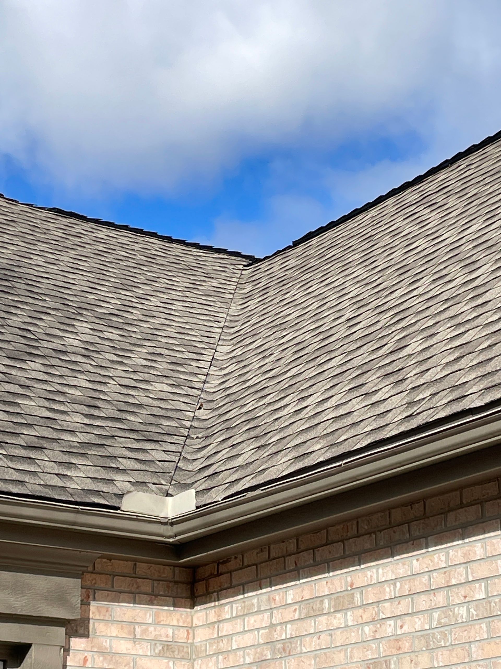 Close-up of a gray shingled roof with brick walls beneath and cloudy blue sky above