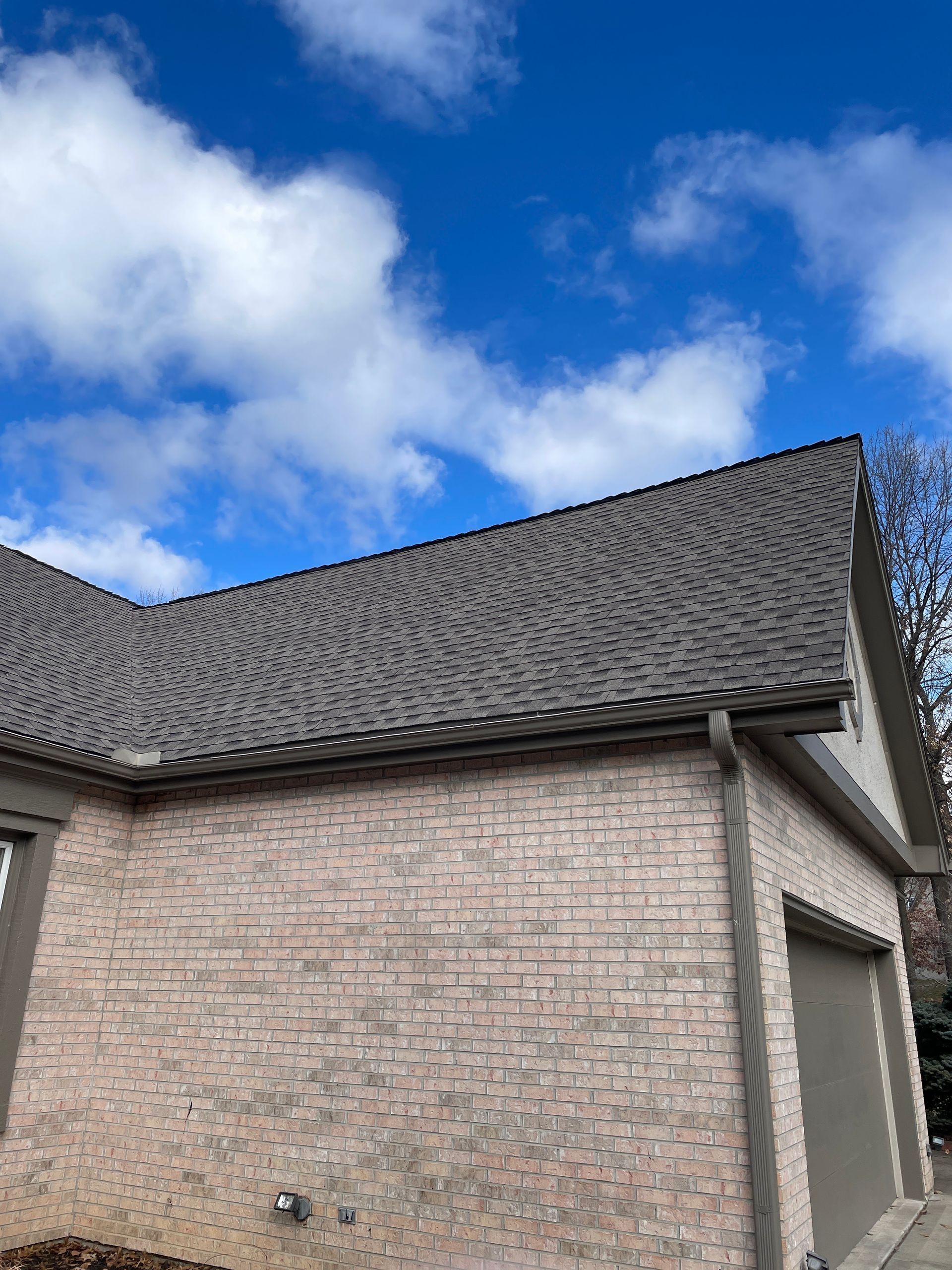 Brick house exterior with gray shingled roof under a blue sky with clouds