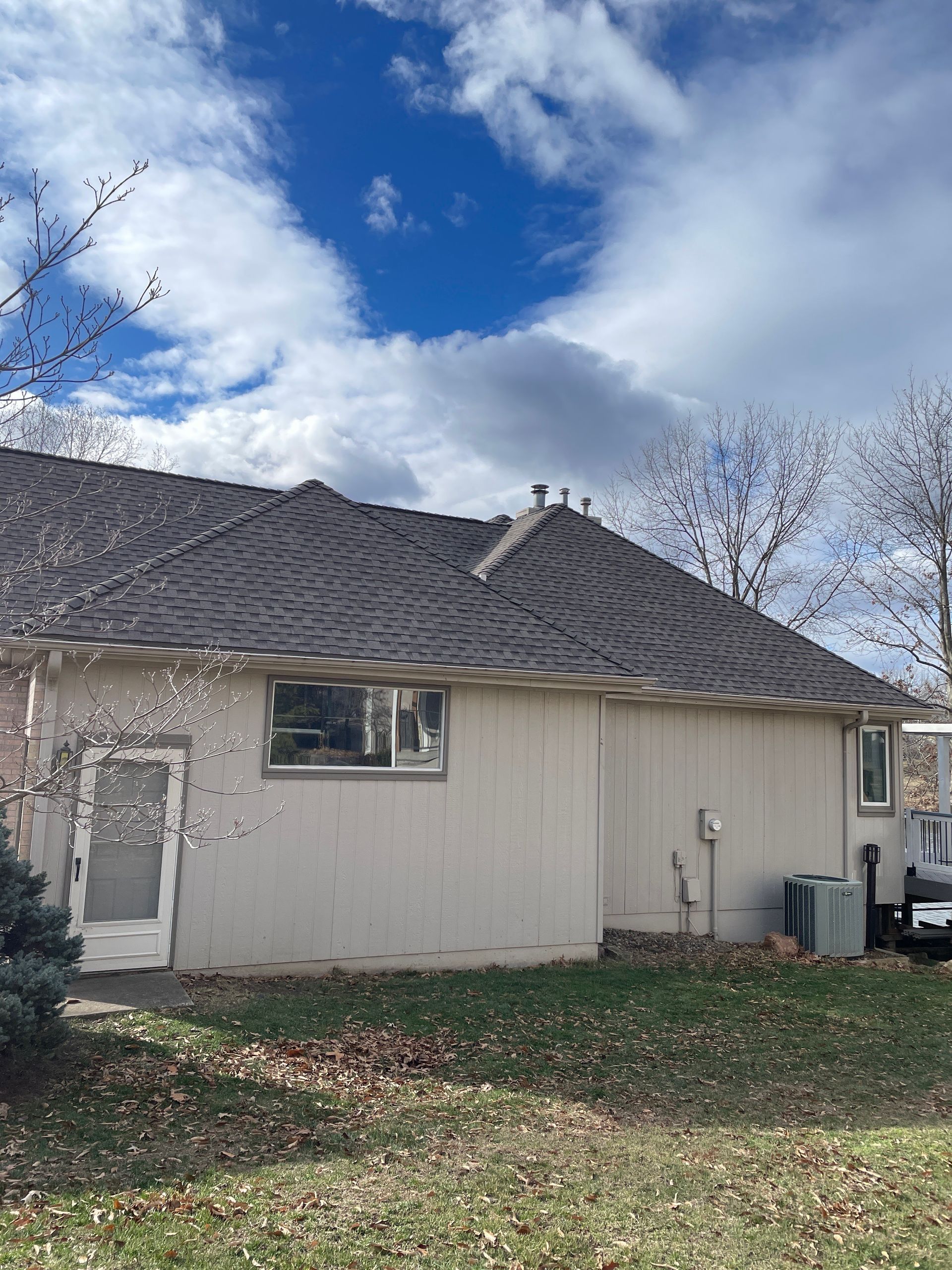 Small beige house with a gray shingled roof under a cloudy blue sky, with trees and fallen leaves in the yard