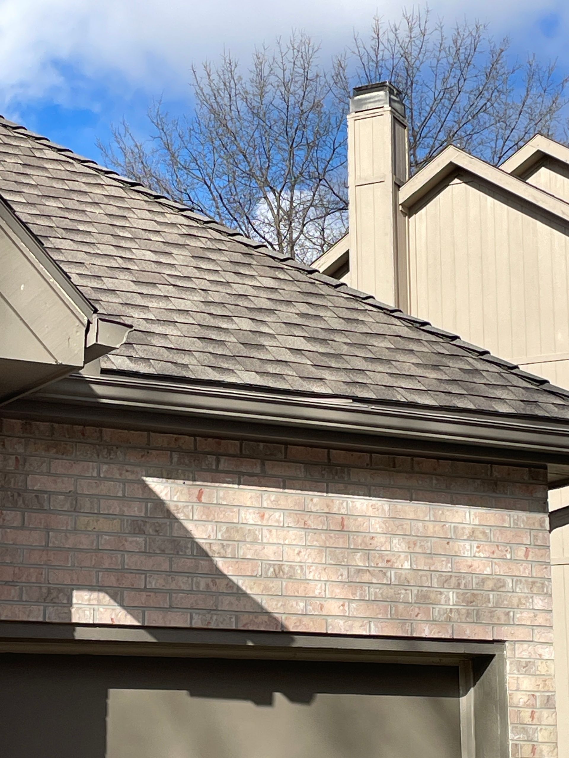 Brick house roof and chimney under a blue sky, with tree branches in the background