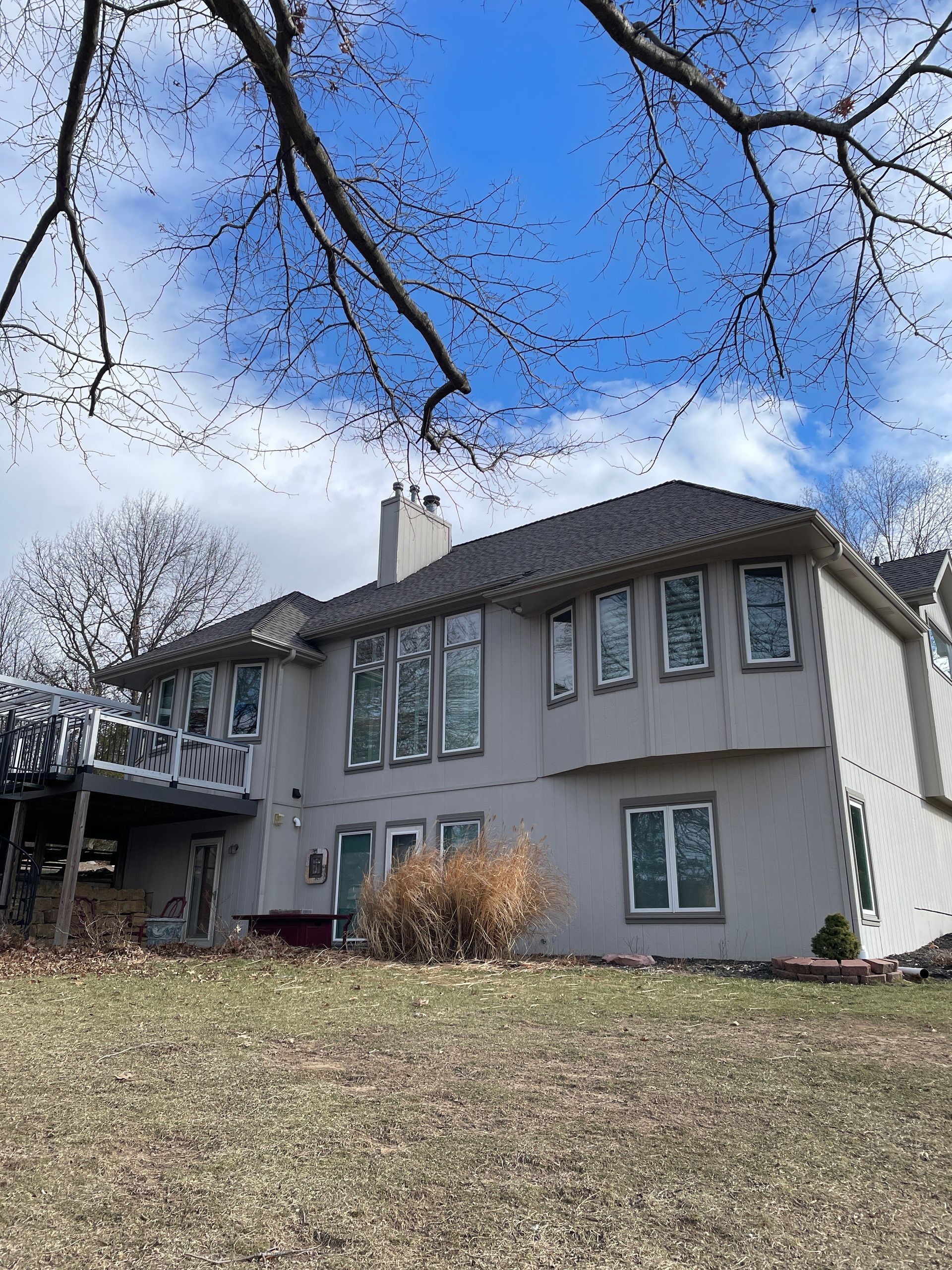 Two-story gray house on a grassy hill under a blue sky with bare tree branches overhead