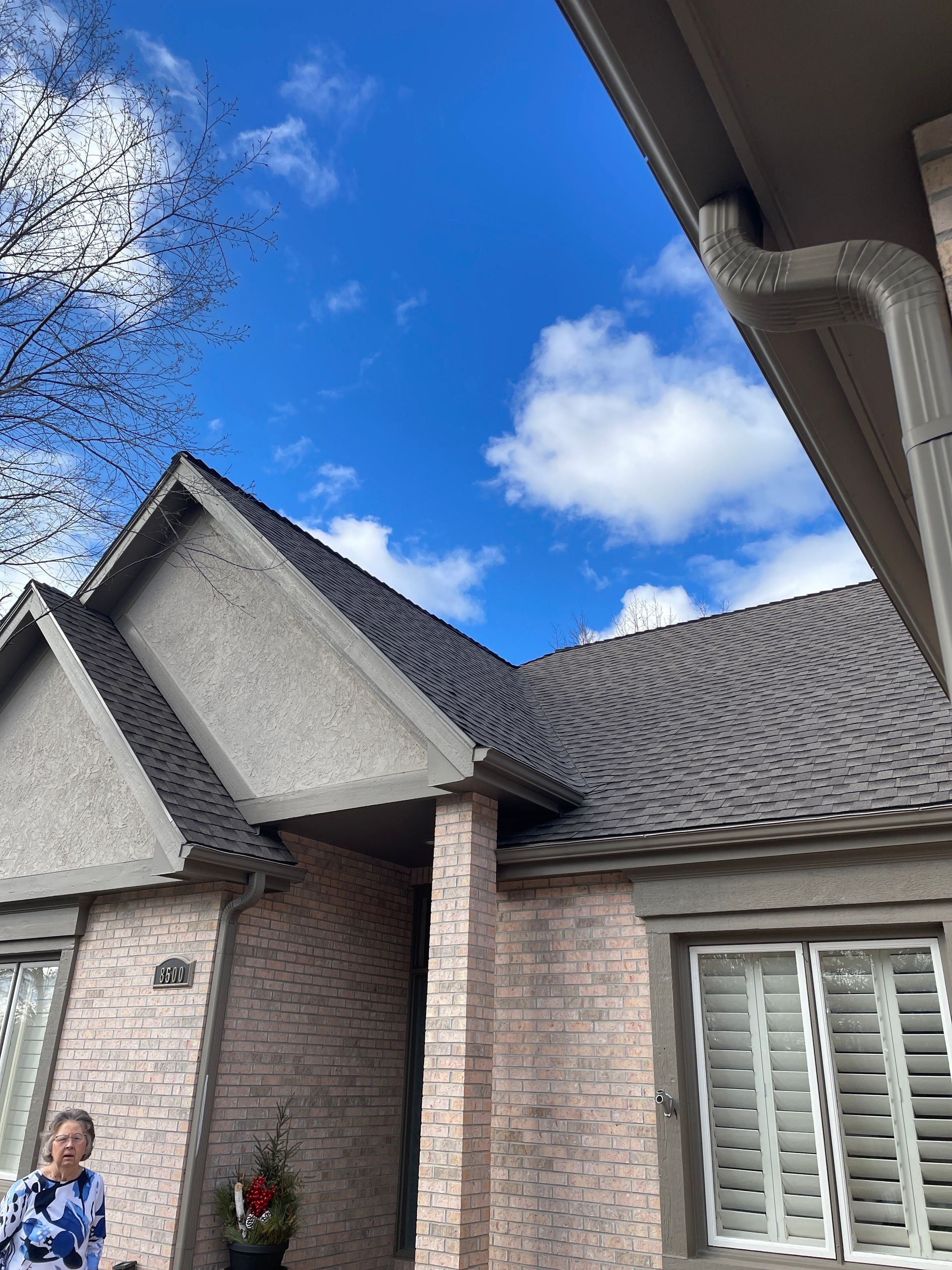 Brick house exterior with gray shingled roof and white shutters under a bright blue sky