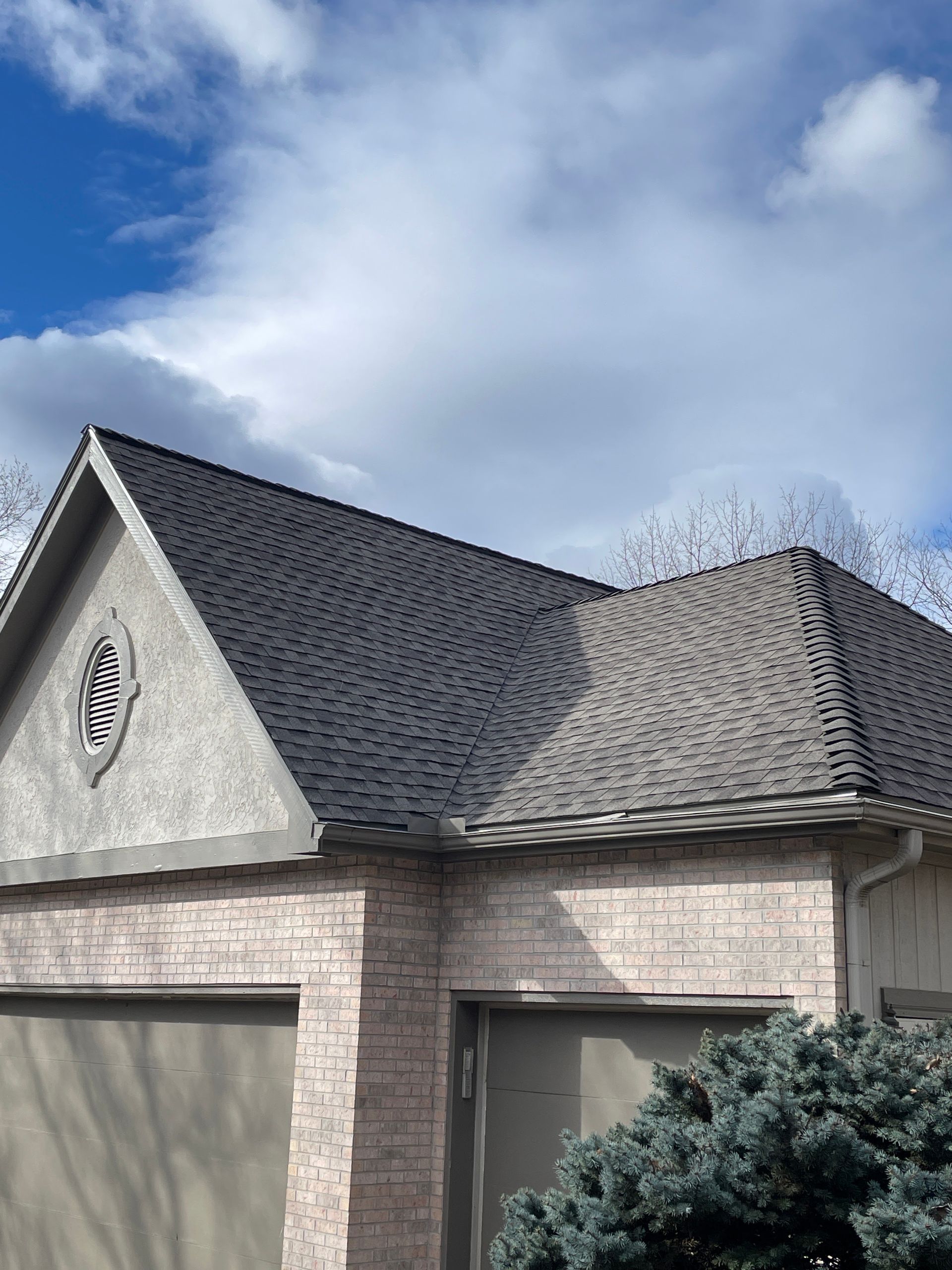 Suburban house roof with gray shingles, stucco walls, and a cloudy blue sky above