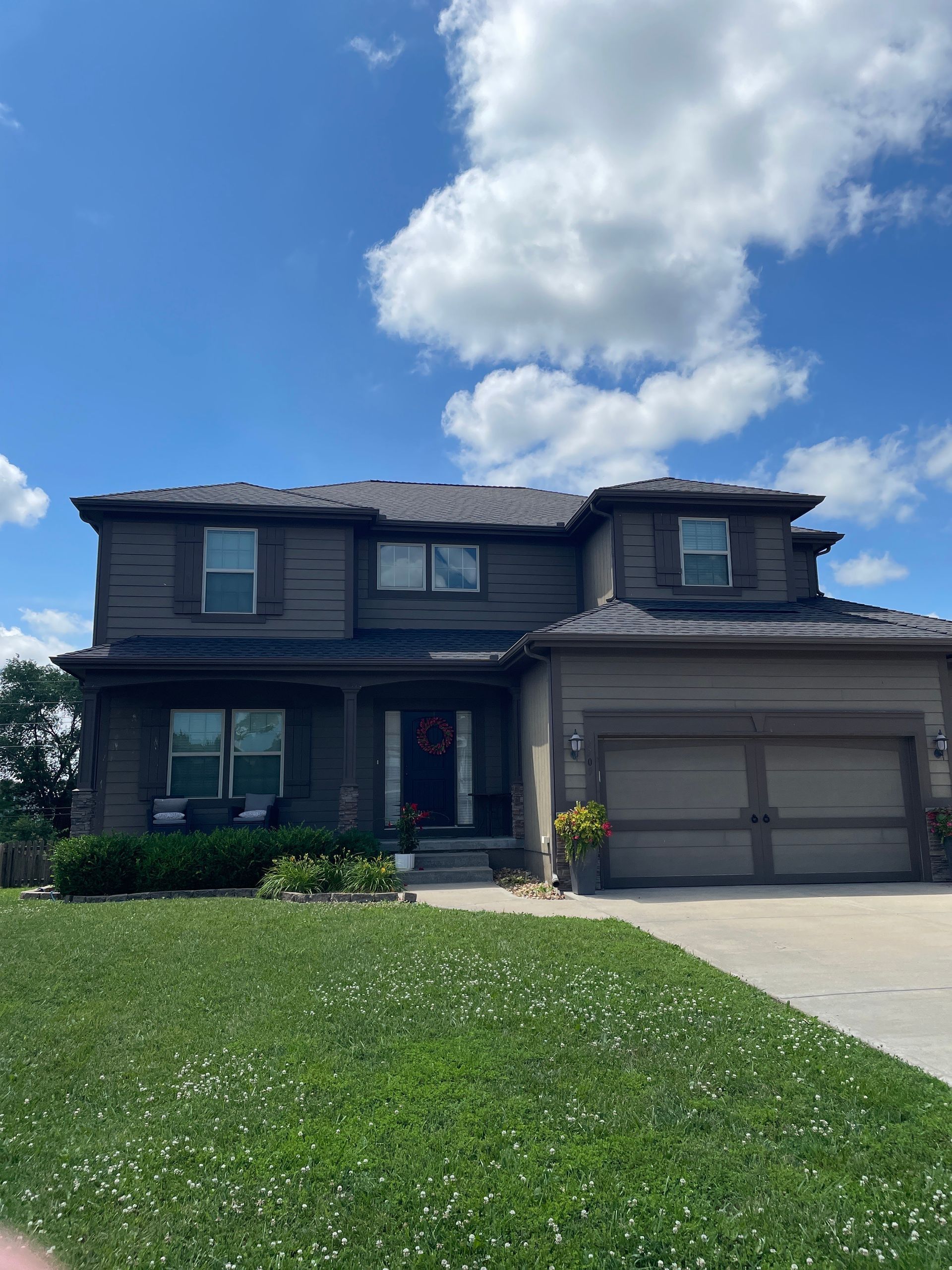 Two-story suburban house with a garage, dark siding, and a green lawn under a bright blue sky.