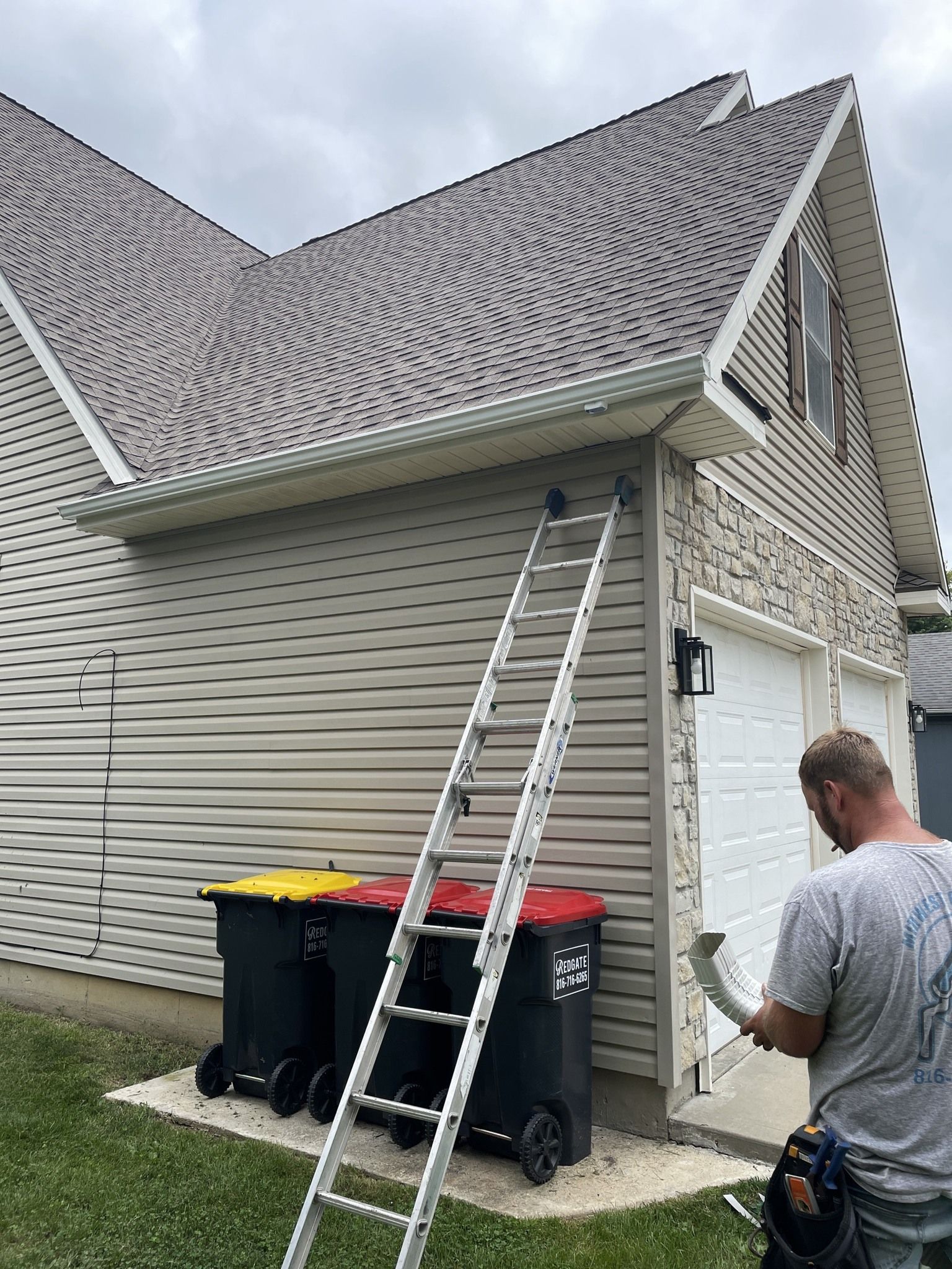 Worker on a ladder inspecting a house roof and siding beside trash bins and a lawn.