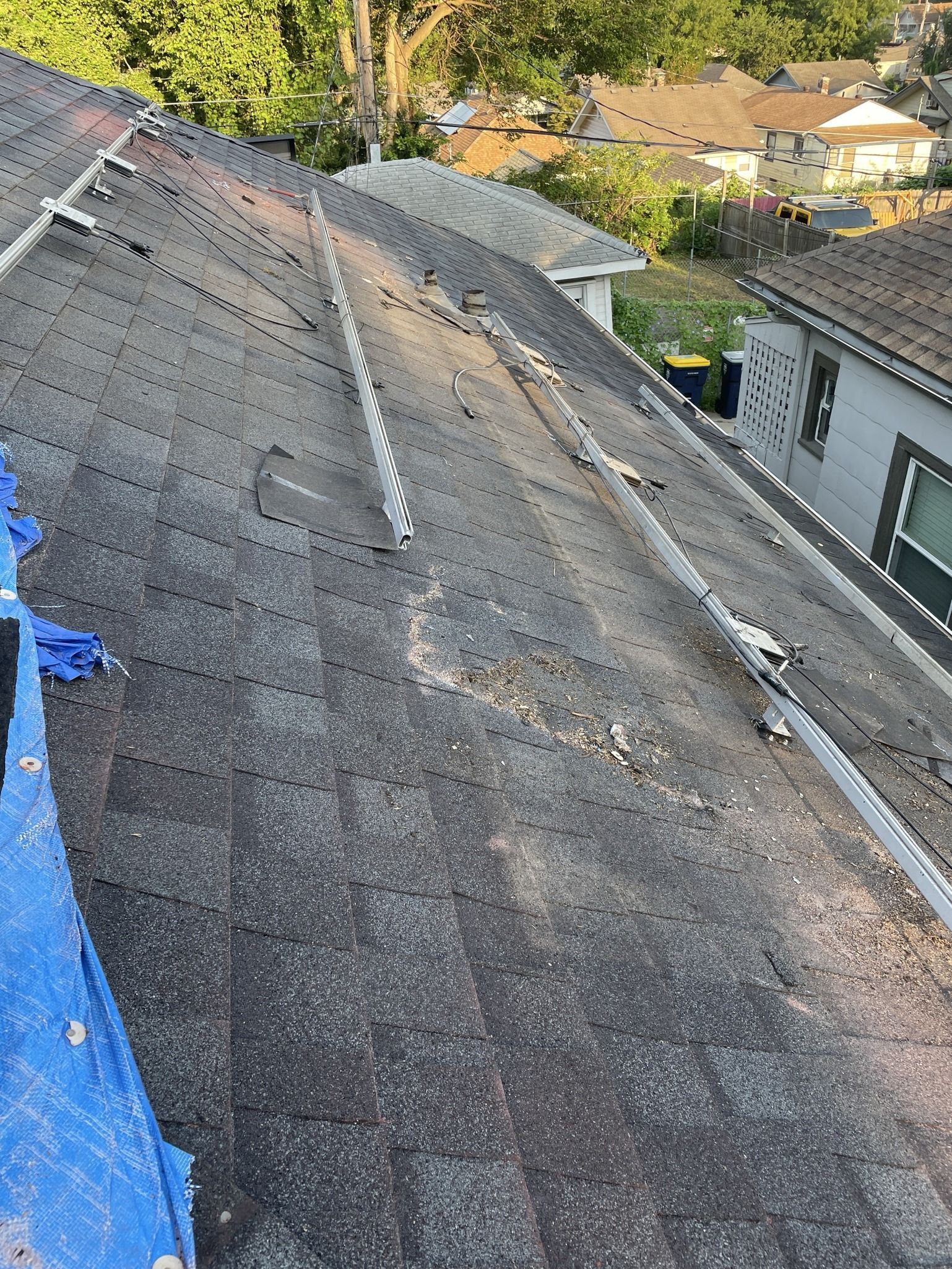 Damaged shingled roof with blue tarp on the left, viewed from above with houses and trees in the background
