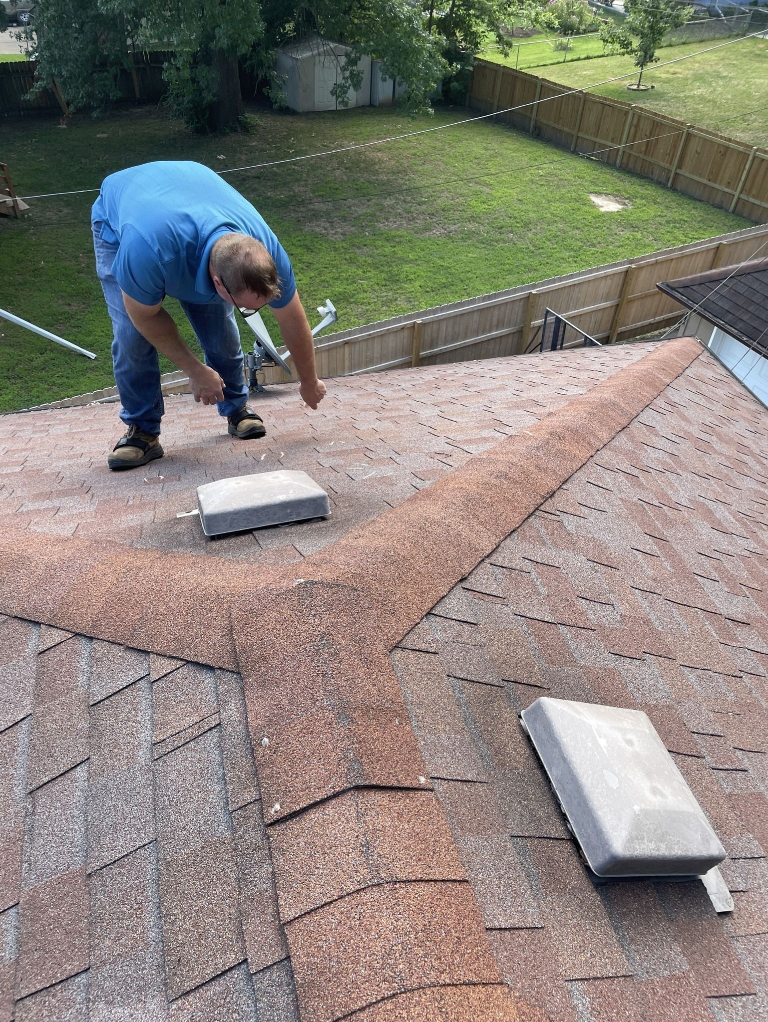 Worker on a shingled roof inspecting and repairing around a skylight in a backyard home.