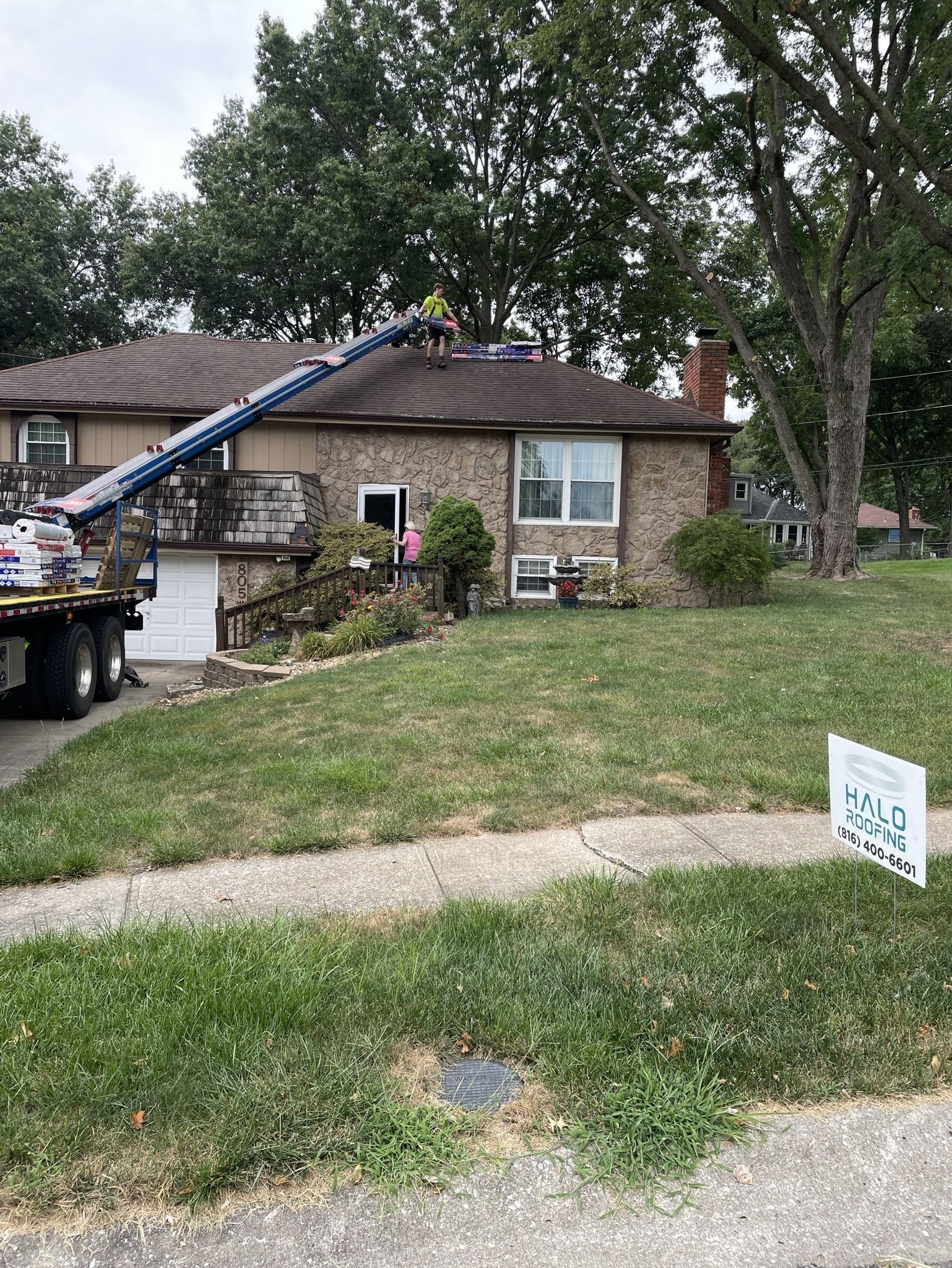 Tree-lined front yard with stone house, utility truck ladder, and a small yard sign in the foreground