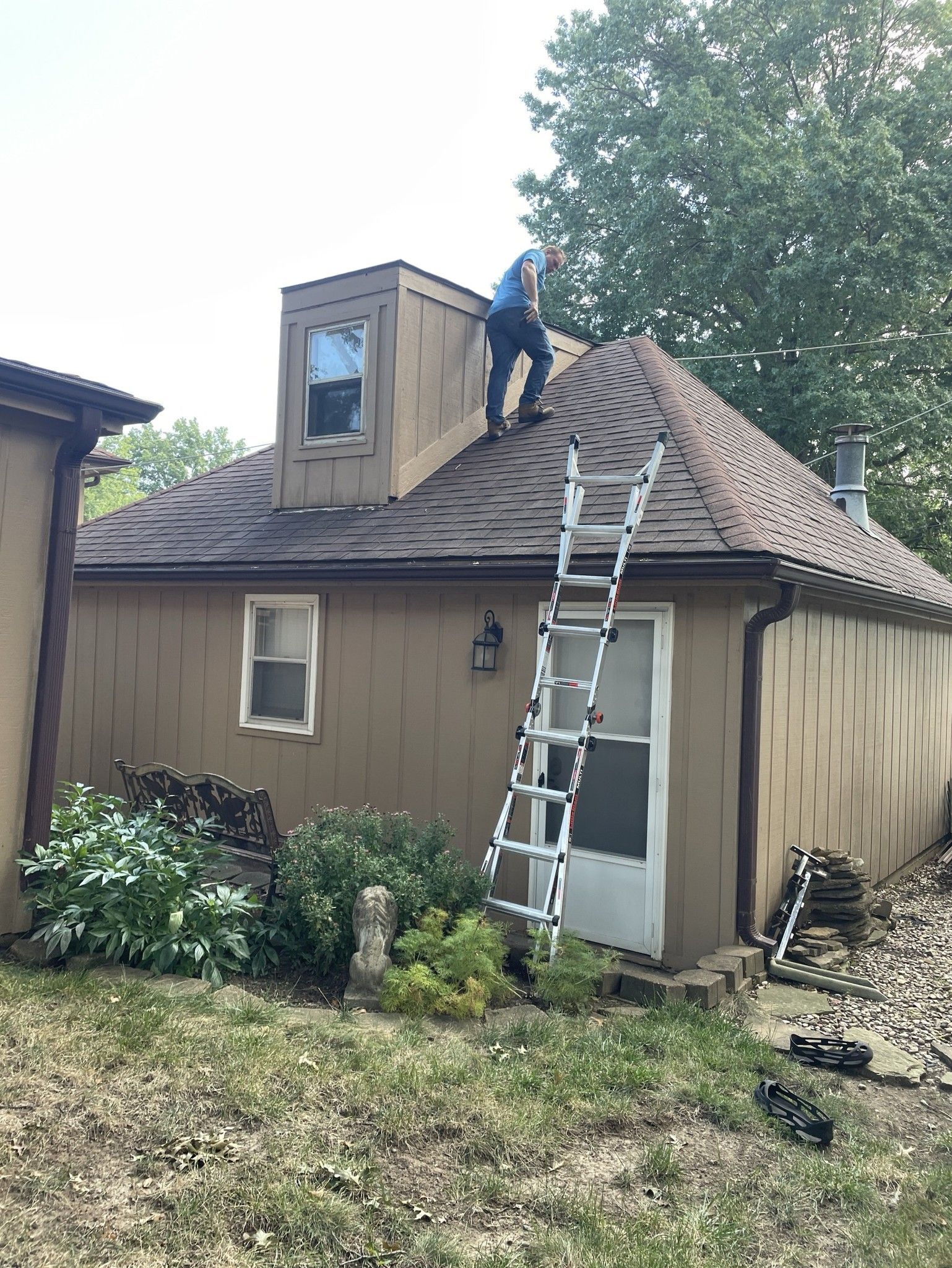 Worker on a roof beside a ladder, repairing shingles on a tan house with a small dormer.