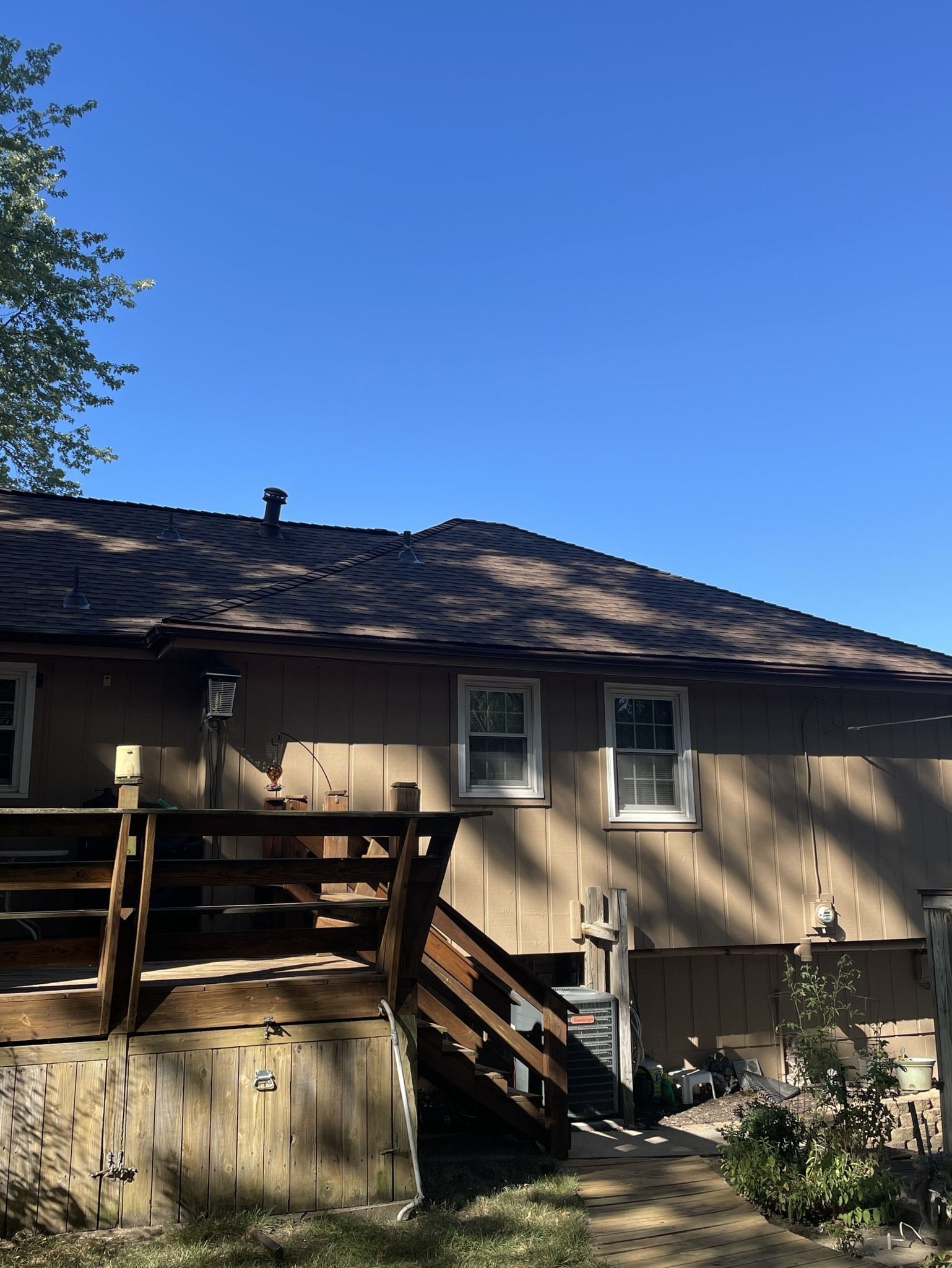 Tan house with a wooden staircase and deck under a blue sky, partially shaded by trees