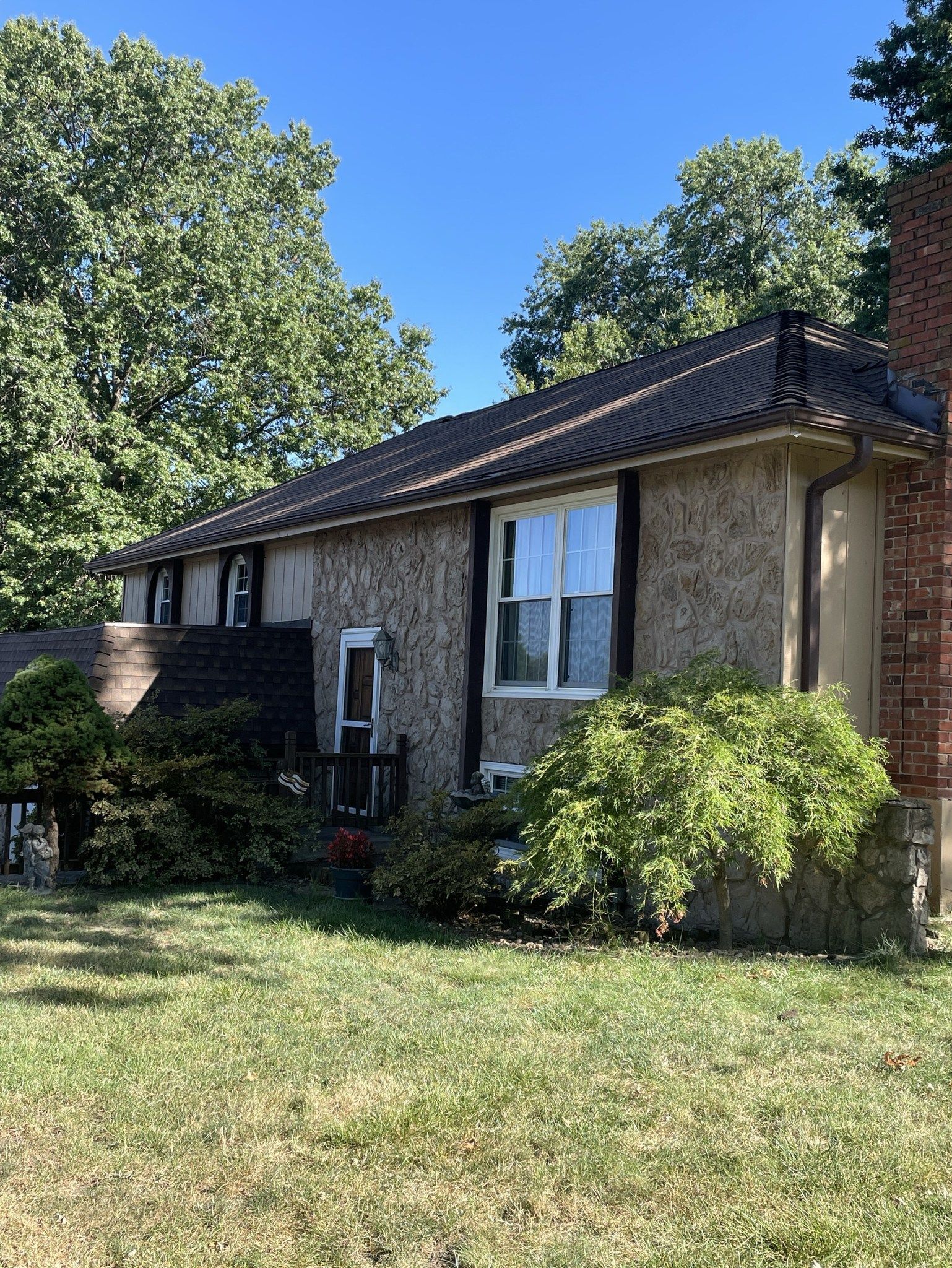 Brick house with dark roof, white window trim, and a green lawn under sunny trees.