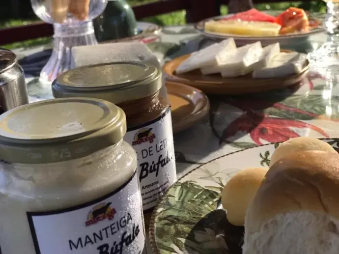 Uma mesa de café da manhã com potes de manteiga de búfala e doce de leite, fatias de queijo branco e pãezinhos.