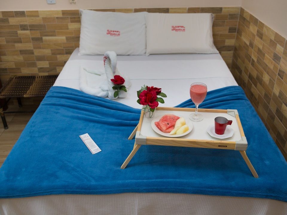 Uma cama com um cobertor azul, uma toalha em forma de cisne, um buquê de rosas vermelhas e uma bandeja com frutas, uma bebida e uma caneca.