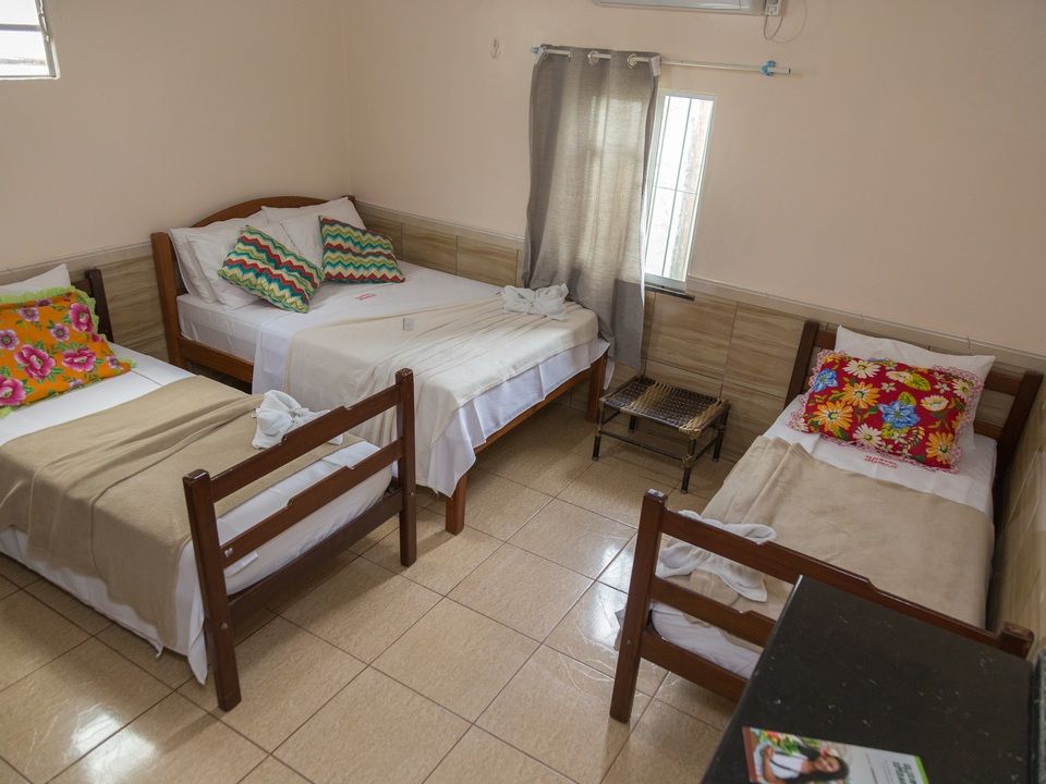 Um quarto com uma cama de casal e duas camas de solteiro, com almofadas estampadas e colchas bege sobre piso de azulejo.