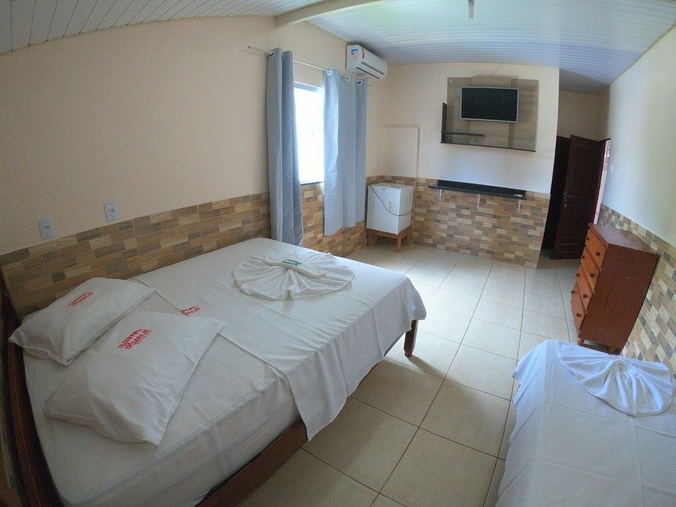 Um quarto de hotel com uma cama de casal, uma cama de solteiro menor, uma cômoda de madeira, uma televisão na parede e piso de azulejo.