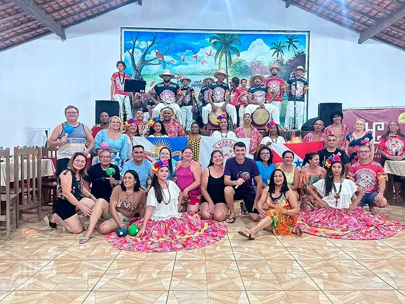 Um grupo com trajes festivos e coloridos posa em um palco de azulejos com um mural ao fundo, alguns segurando instrumentos e adereços.