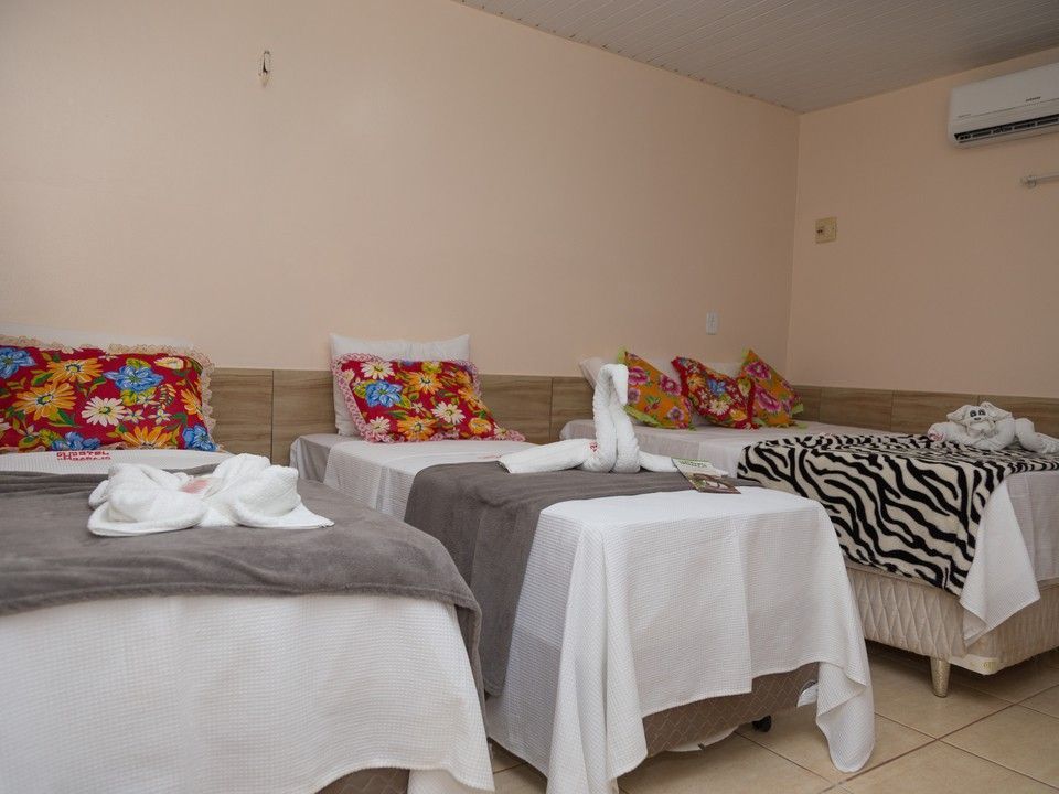 Um quarto de hotel com três camas de solteiro, cada uma com lençóis brancos, caminhos de mesa cinza, travesseiros coloridos e toalhas em formato de cisne.