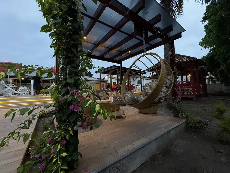 Um pátio externo com pérgola, cadeira suspensa de vime, trepadeiras floridas roxas e um gazebo vermelho nas proximidades.