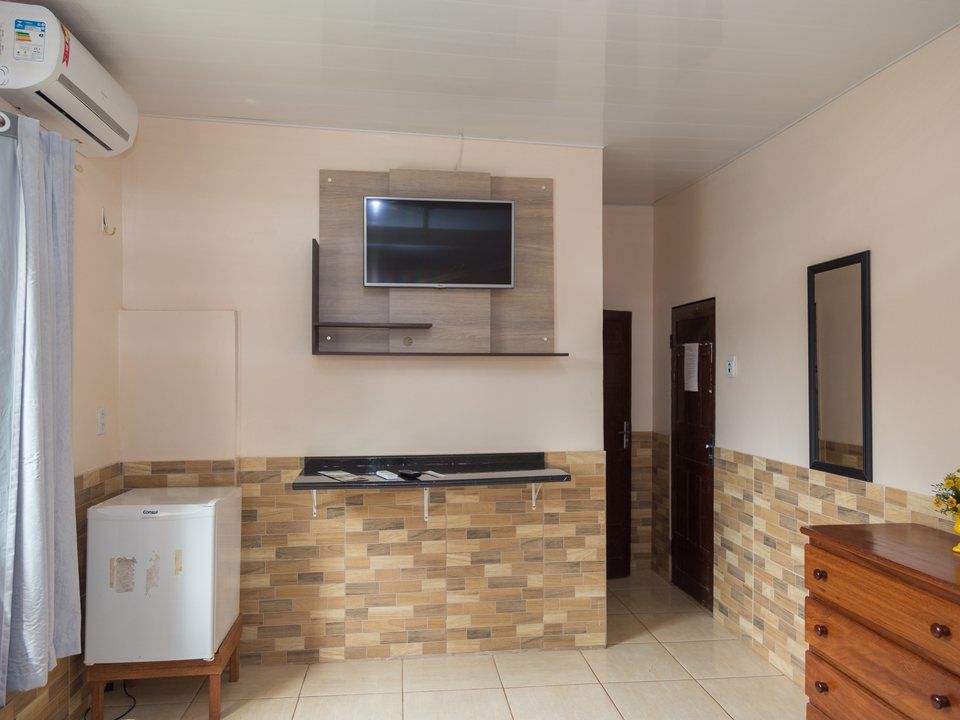 Um quarto mobiliado com paredes claras, uma pequena geladeira branca, TV fixada na parede, prateleiras, cômoda de madeira e uma porta.