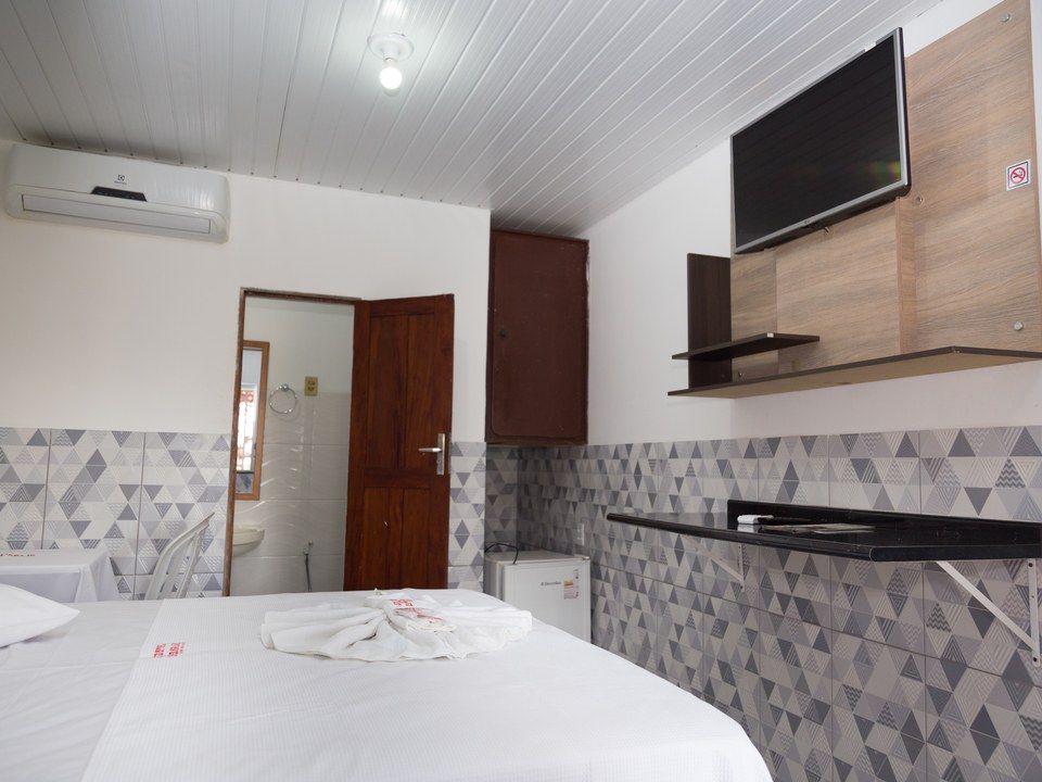 Um pequeno quarto de hotel com uma cama, uma televisão fixada na parede, uma escrivaninha também fixada na parede e uma porta que dá acesso a um banheiro.