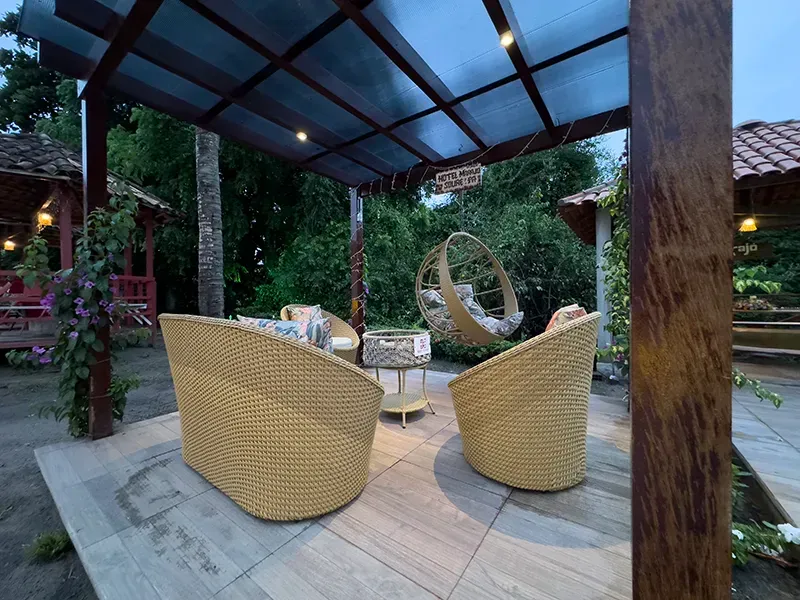 Um pátio externo com cadeiras de vime bege, uma mesa lateral combinando e uma cadeira de balanço suspensa sob uma pérgola de madeira escura.