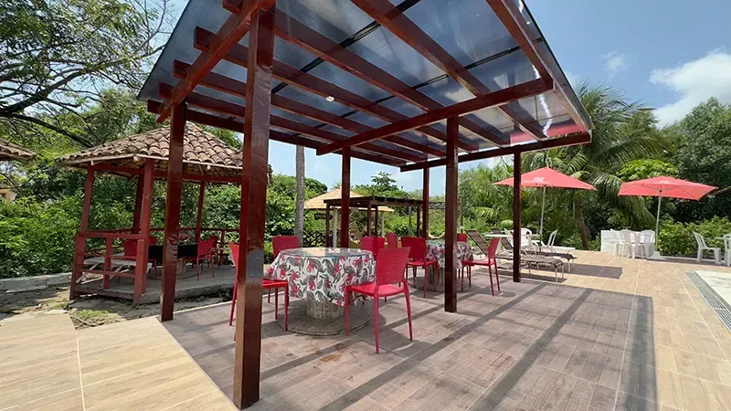 Pátio externo com pérgola de madeira, mesas estampadas com cadeiras vermelhas e guarda-sóis vermelhos junto à piscina.