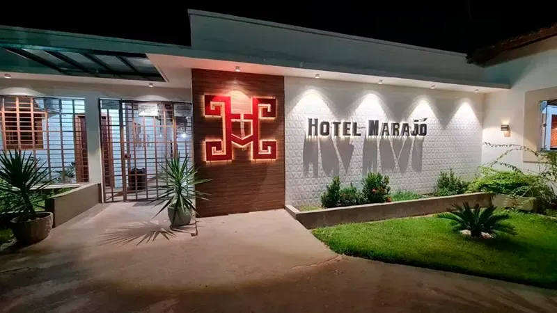 Vista exterior do Hotel Marajó à noite, com a placa e o logotipo iluminados numa parede de pedra branca e paisagismo em frente.