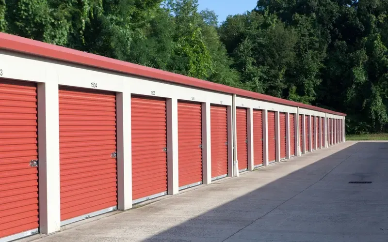 Row of red storage unit doors along a paved lane beside trees