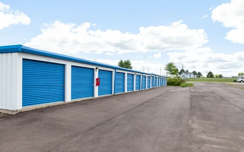 Row of blue storage unit doors beside an empty paved lot under a partly cloudy sky