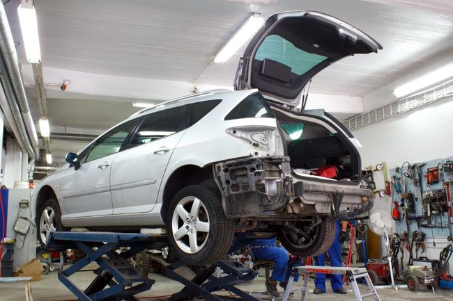 Silver car raised on a lift in a garage, with the rear bumper removed and the trunk open, tools on wall in the background.