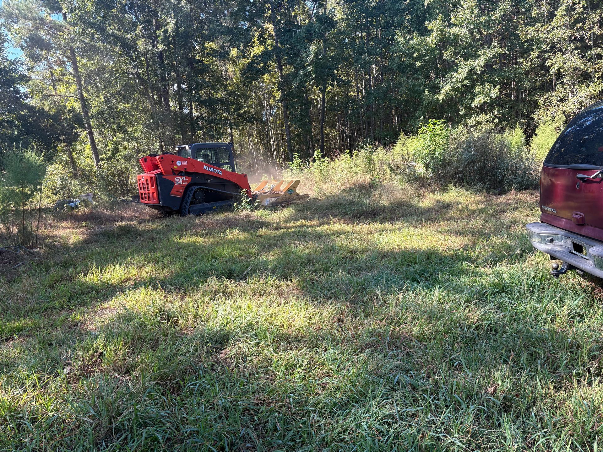 Red skid steer clearing brush in a grassy field with trees in the background. A red truck is parked on the right.