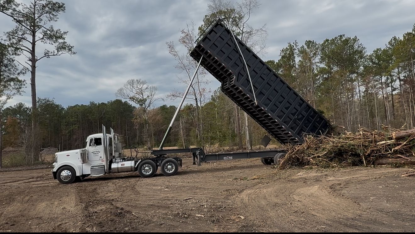A dump truck dumping debris in a wooded area under a cloudy sky.