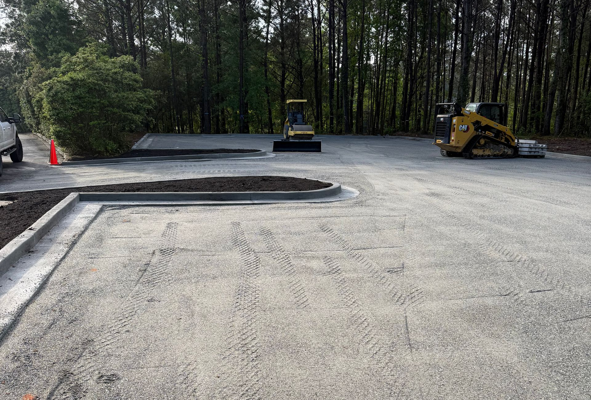 Gravel parking area under construction with concrete curbing, two small excavators, and trees in the background.